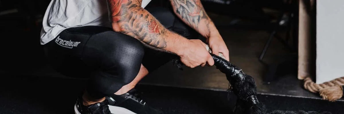 An image of a heavily tattooed person kneeling down, wearing Bracelayer's compression leggings with knee support.